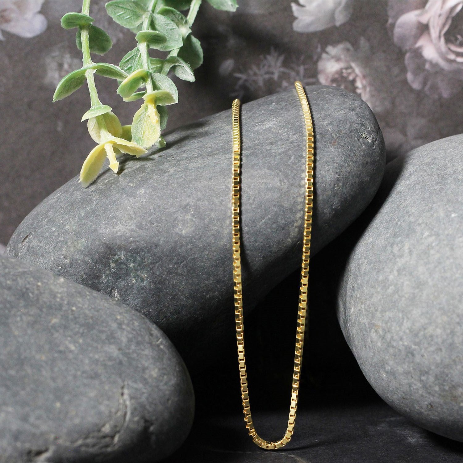 A 14K yellow gold box chain draped over smooth gray stones.