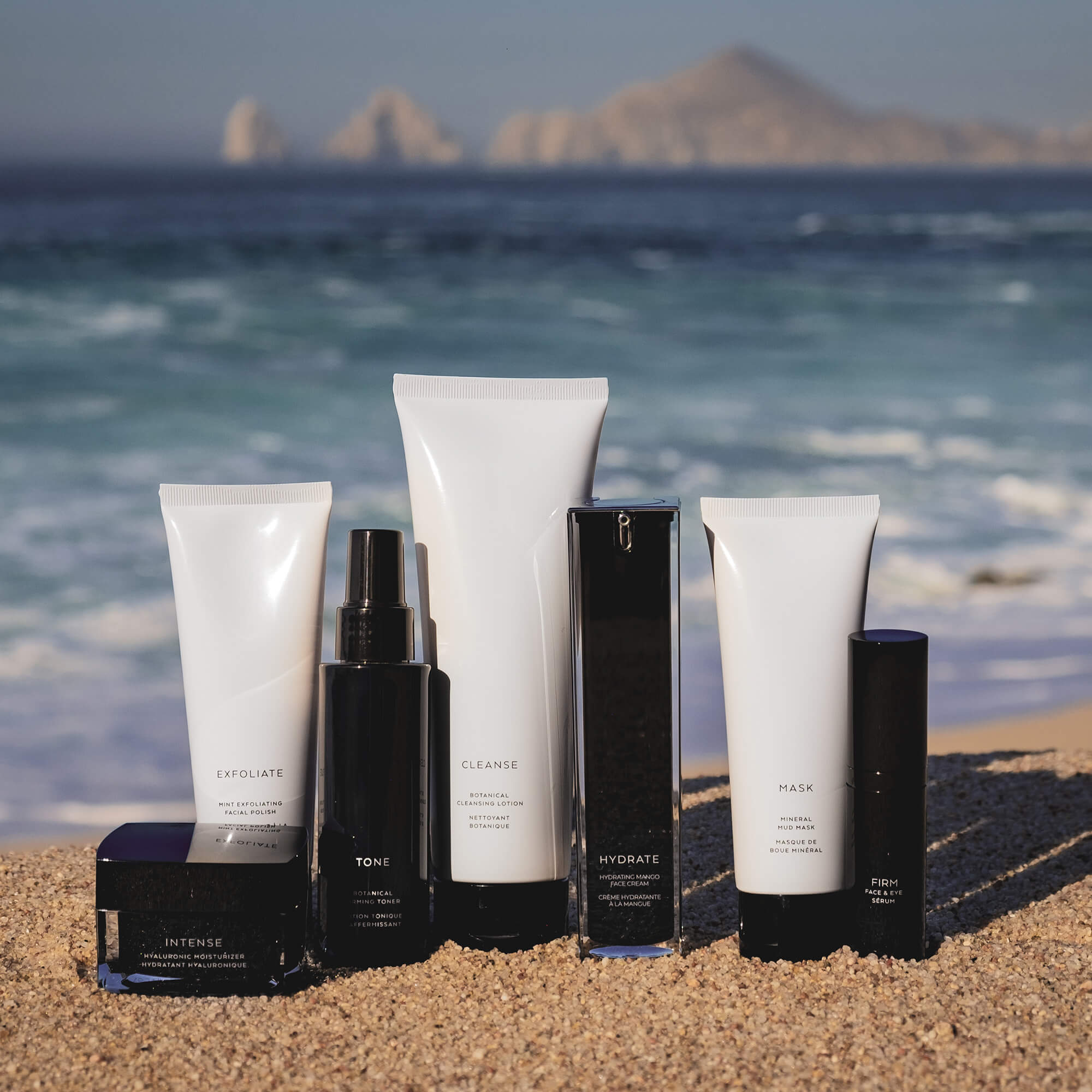A collection of skincare products, including Firm Serum, on a sandy beach with the ocean and mountains in the background.