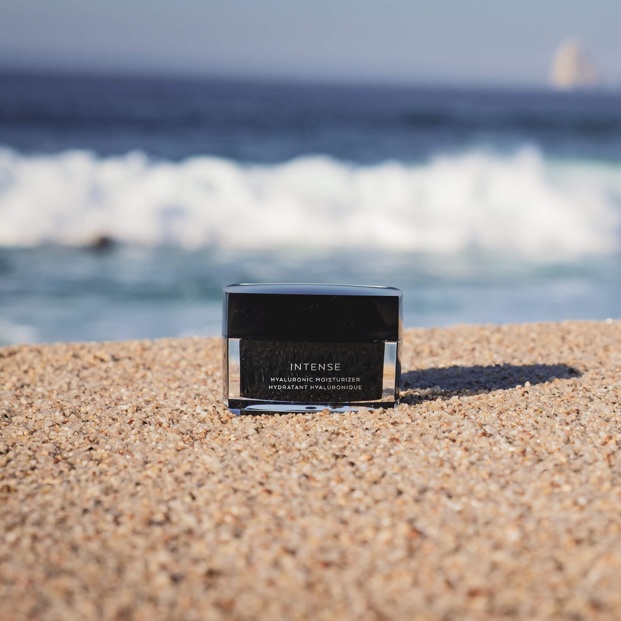 Black jar of "Intense Hyaluronic Moisturizer" on a sandy beach with ocean waves.