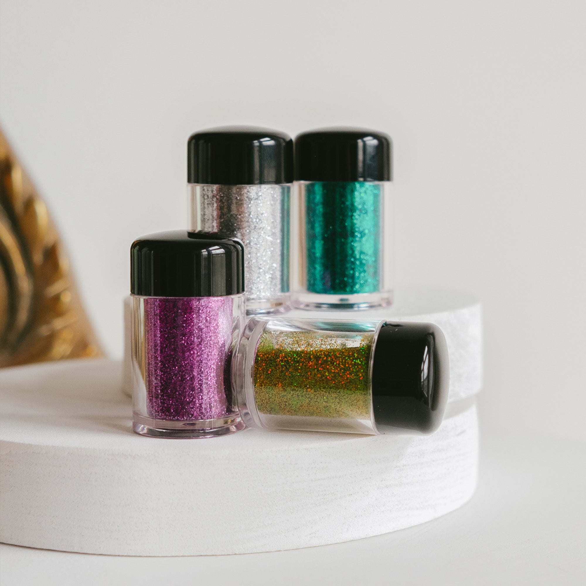 Four jars of glitter in purple, silver, green, and gold are displayed on a white surface.
