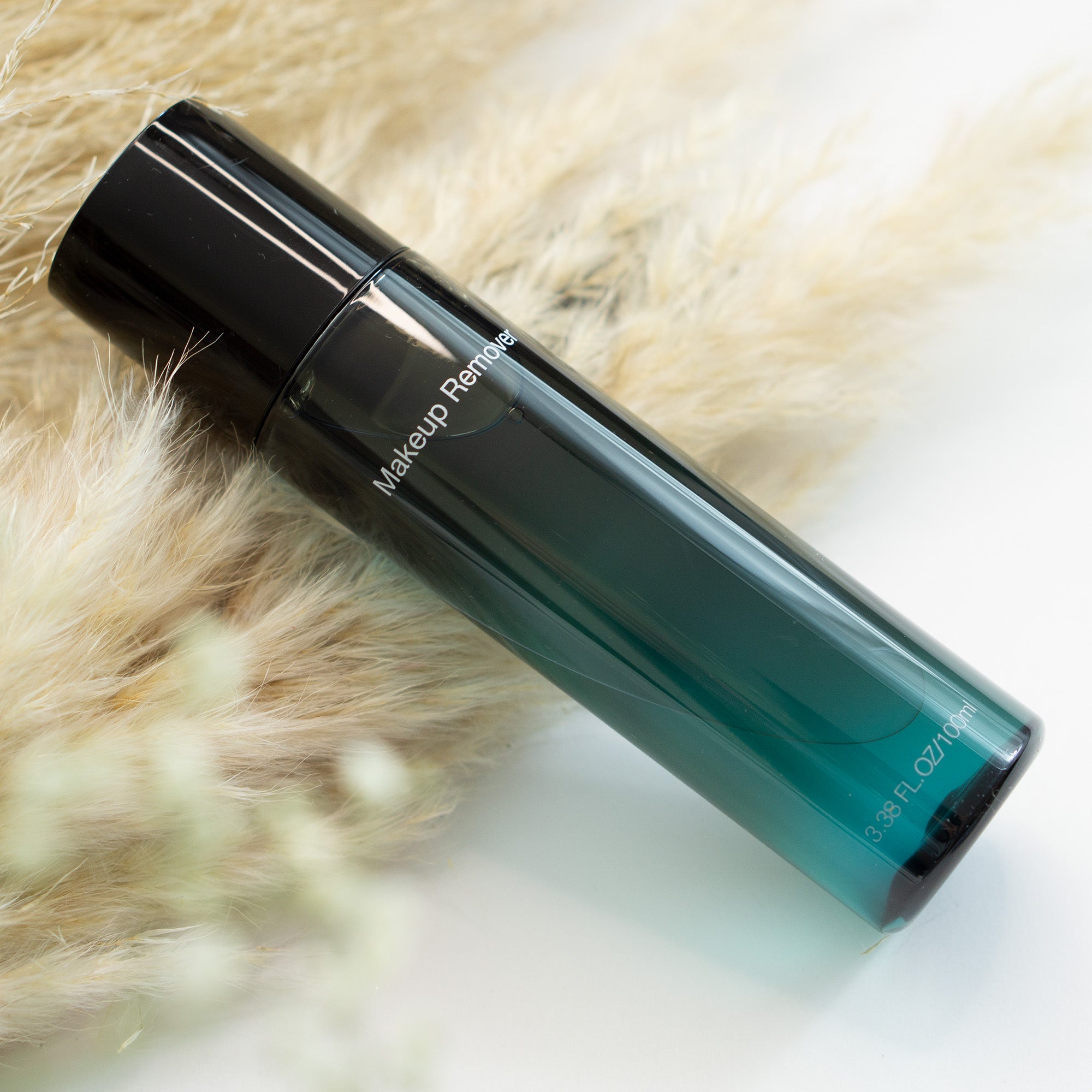 A black and teal makeup remover bottle lies on a bed of dried pampas grass.