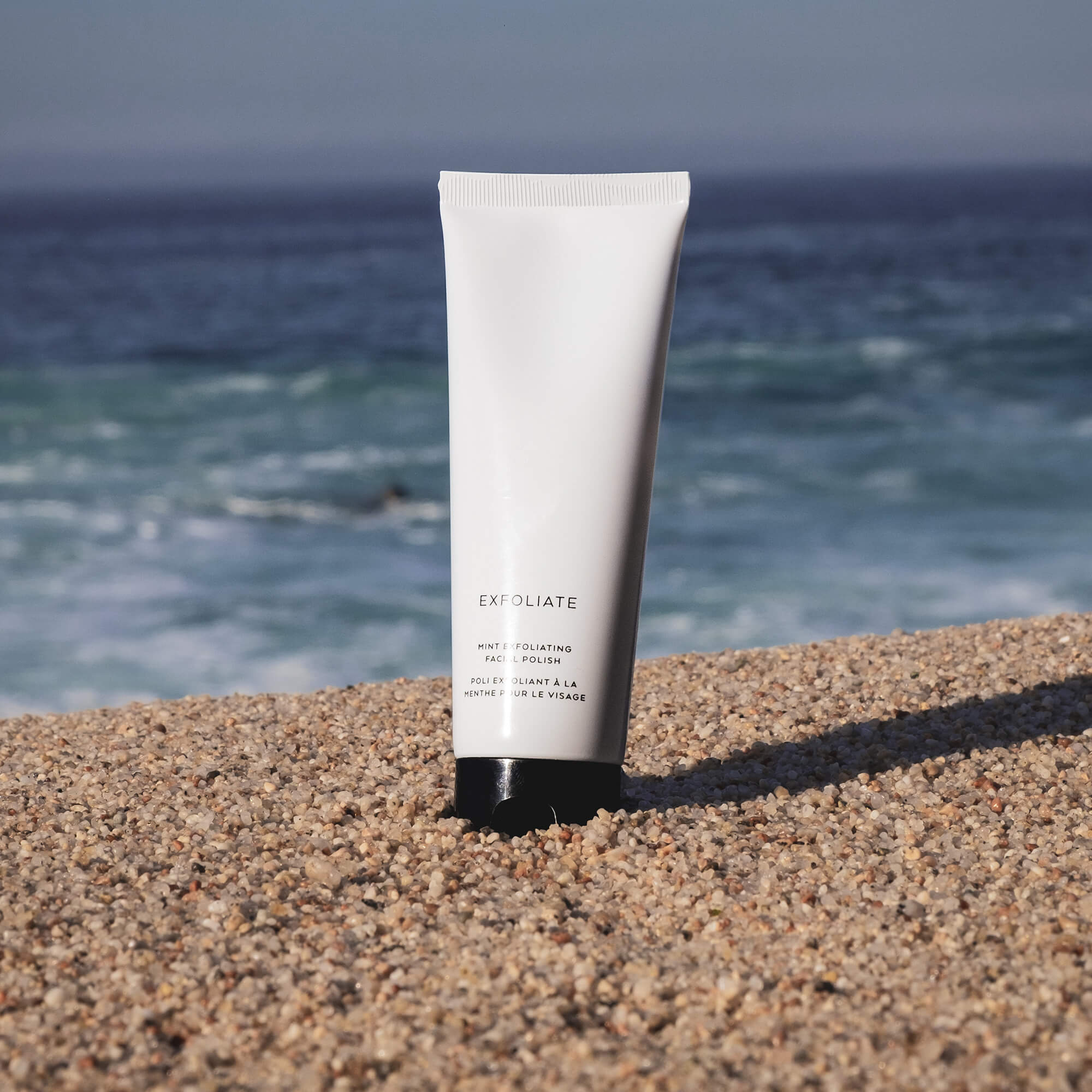 White tube of Mint Exfoliating Facial Polish on a sandy beach with ocean in background.
