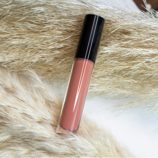 A tube of peach-colored lip gloss with a black cap rests on dried pampas grass.