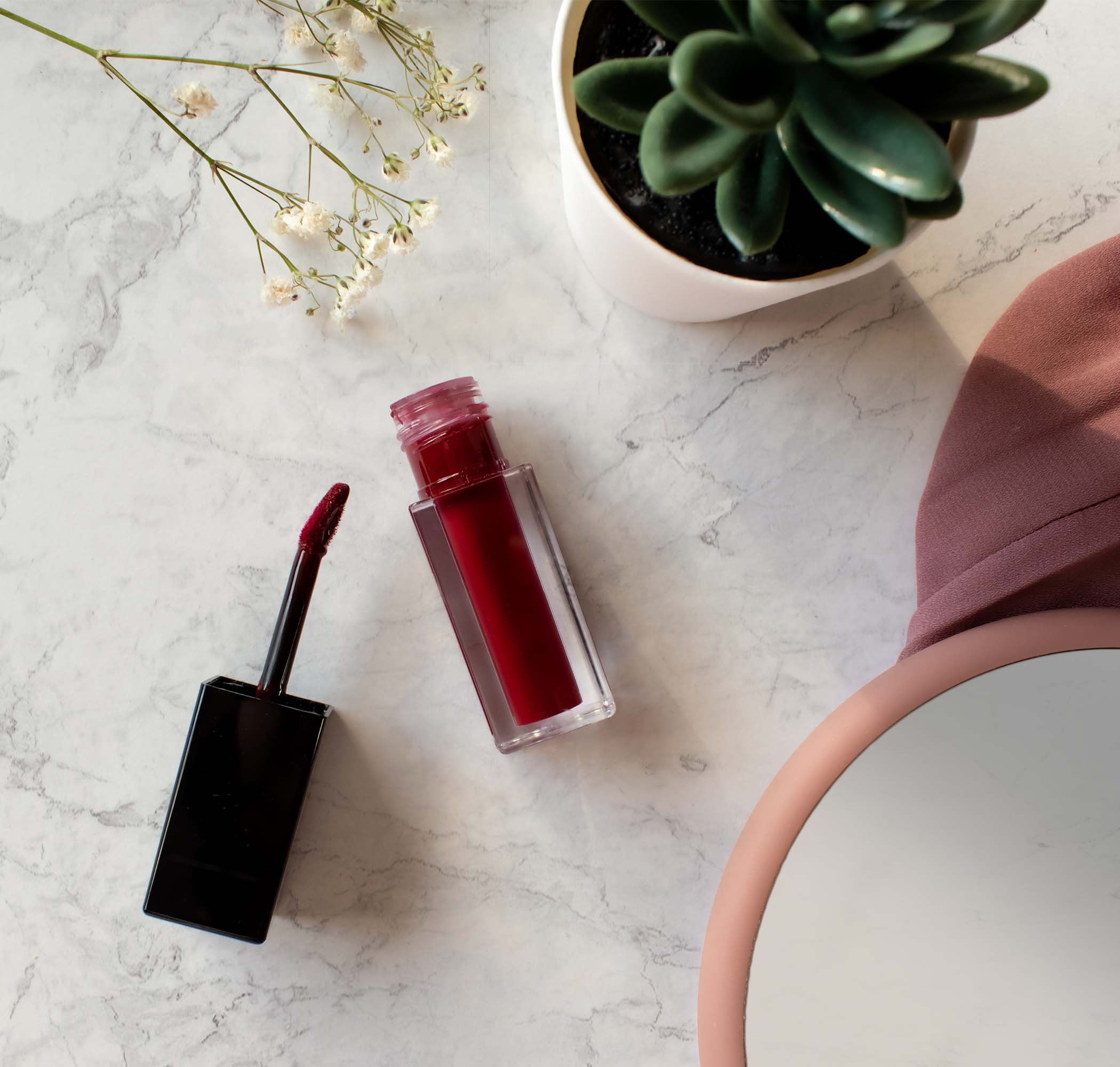 A dark cherry matte lip stain with its applicator on a marble surface with a plant and flowers.