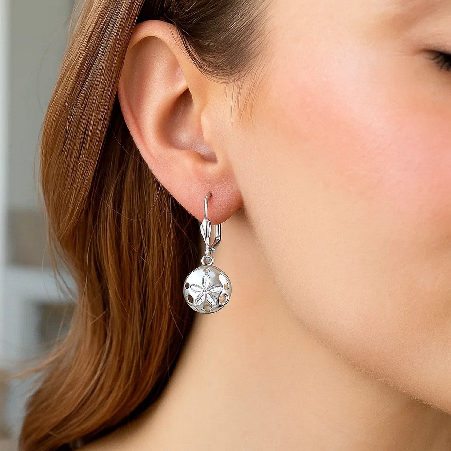 Sterling silver sand dollar dangle earrings with a polished finish are worn on a woman's ear.