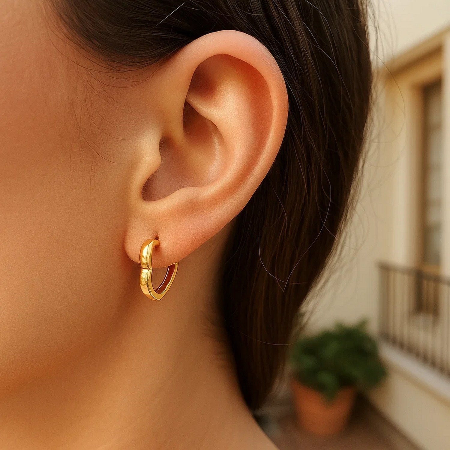 A woman wears a single 14k yellow gold heart huggie hoop earring.