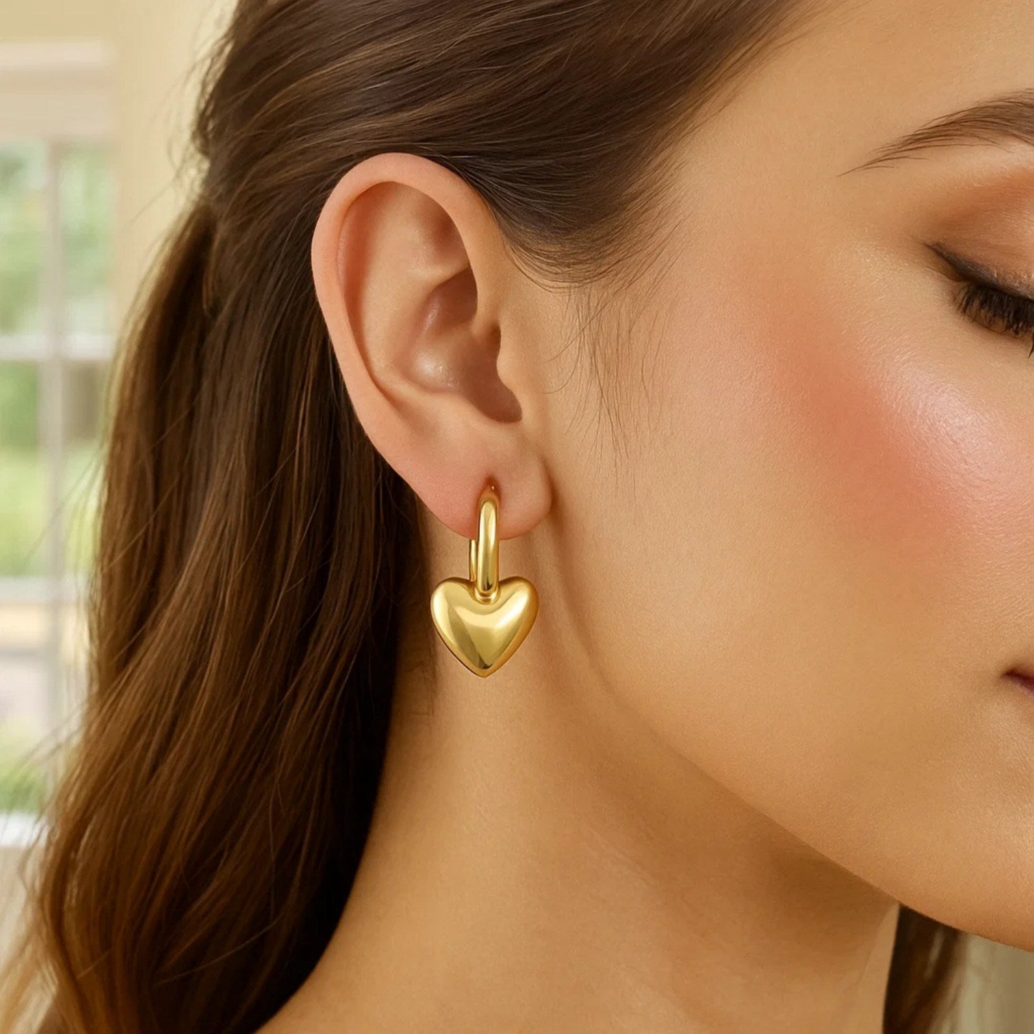 A woman wears a 14K yellow gold puffy heart dangle huggie hoop earring.