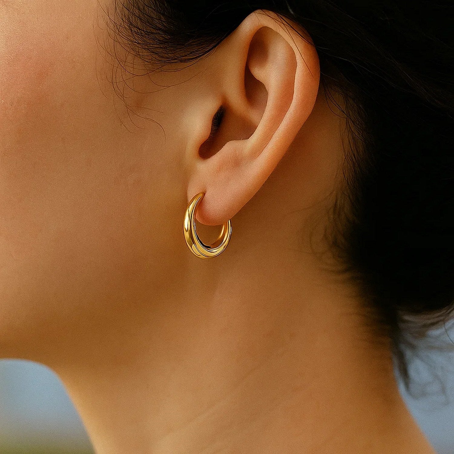 A woman wears a single 14k yellow gold oval hoop earring.