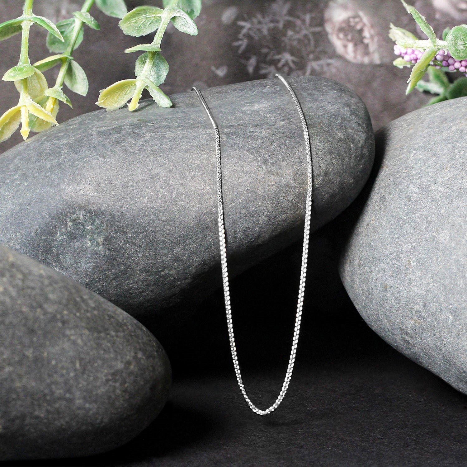 A 14k white gold round wheat chain is draped over smooth grey stones.
