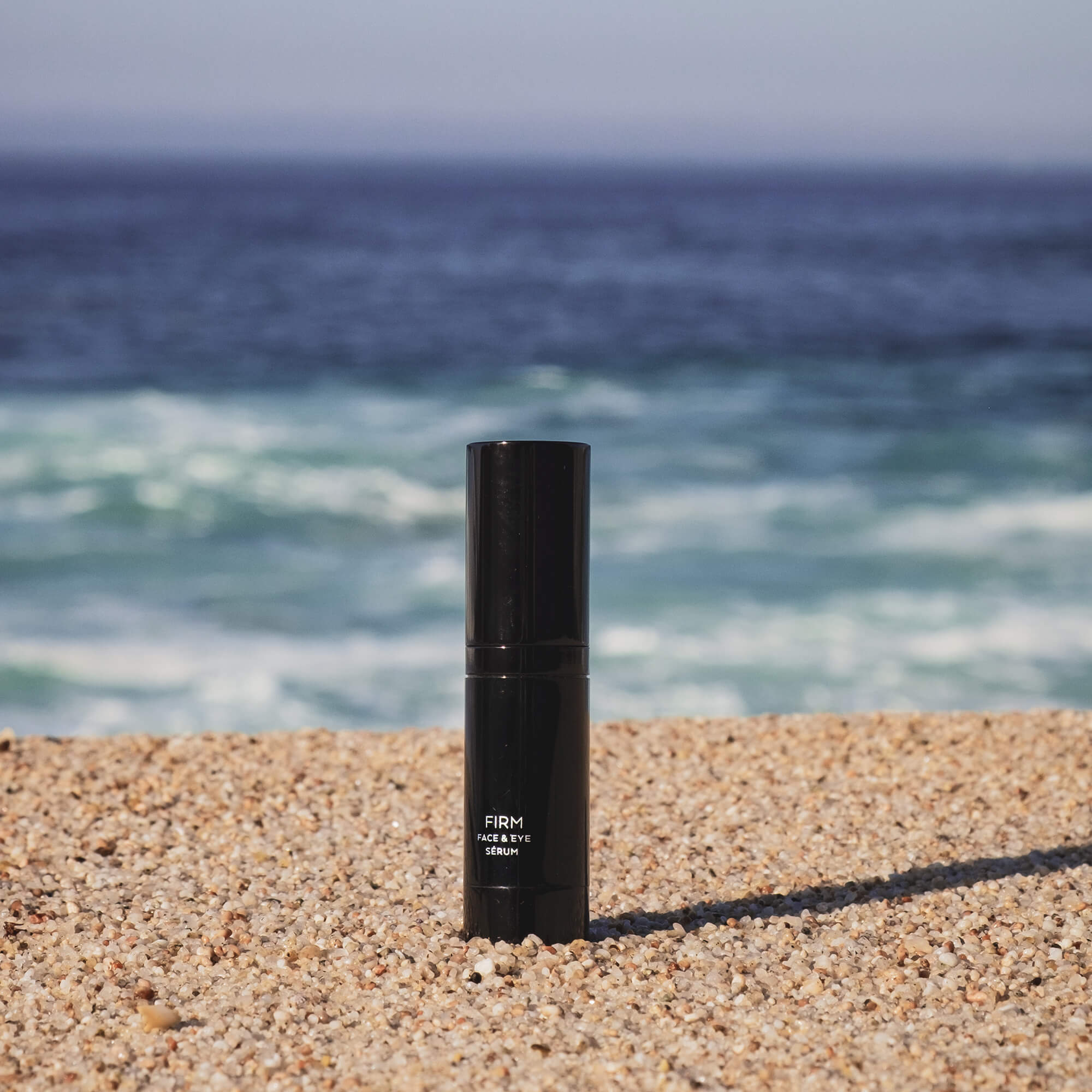 Black Firm Face & Eye Serum bottle on a sandy beach with ocean in the background.