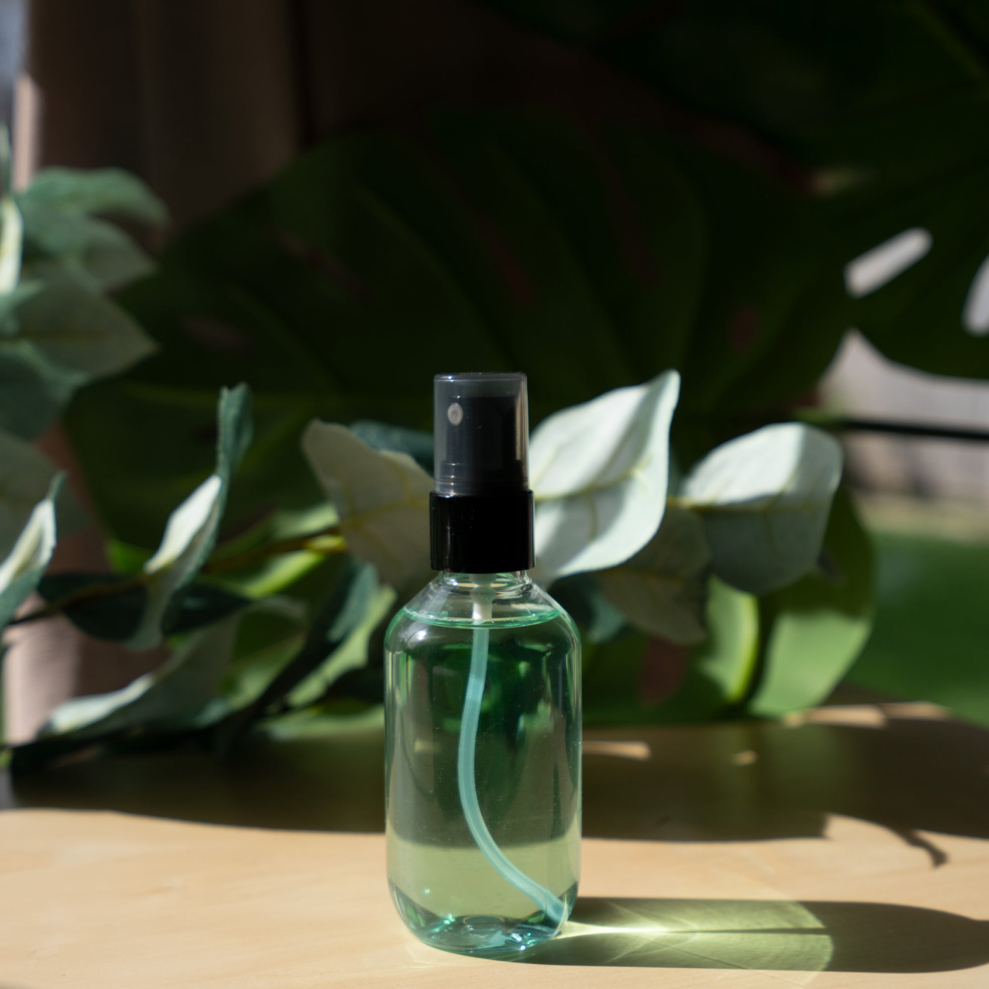 A clear spray bottle with green liquid stands on a wooden surface with green leaves.